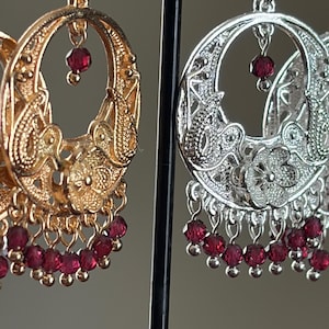 Op de afbeelding: Gouden en zilveren filigraan oorringen met bungelende rode kralen. De oorbellen hebben ingewikkelde bloemen- en paisley-ontwerpen. De gouden oorring staat links en de zilveren oorring staat rechts.