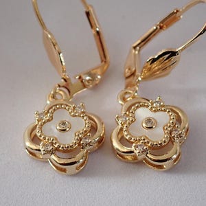 Peut inclure: Une paire de boucles d'oreilles dorées à fermoir. Chaque boucle d'oreille présente un motif de trèfle à quatre feuilles avec un centre blanc et des cristaux clairs étincelants. Les boucles d'oreilles sont conçues avec une fermeture à levier.