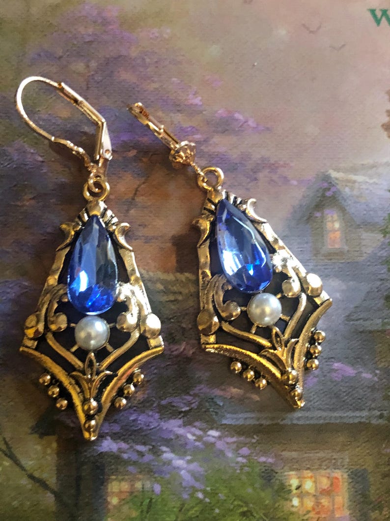 Op de afbeelding: Een paar sierlijke, goudkleurige oorbellen. Elke oorbel heeft een grote, druppelvormige blauwe edelsteen, een kleine parel en ingewikkelde filigraan details. Ze hebben een beugel sluiting en een vintage uitstraling.