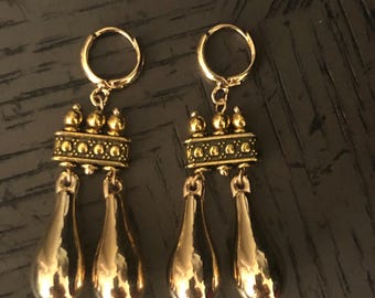 May include: A pair of gold-colored earrings. Each earring features a small hoop at the top, connected to a decorative bar with small, round accents. Below the bar hang three teardrop-shaped pendants. The earrings are displayed against a dark background.