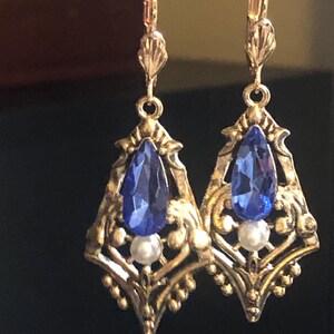 Op de afbeelding: Een paar goudkleurige filigraan oorbellen met een druppelvormige blauwe edelsteen en een kleine witte parel. De oorbellen hebben een beugel sluiting en een decoratief ontwerp.