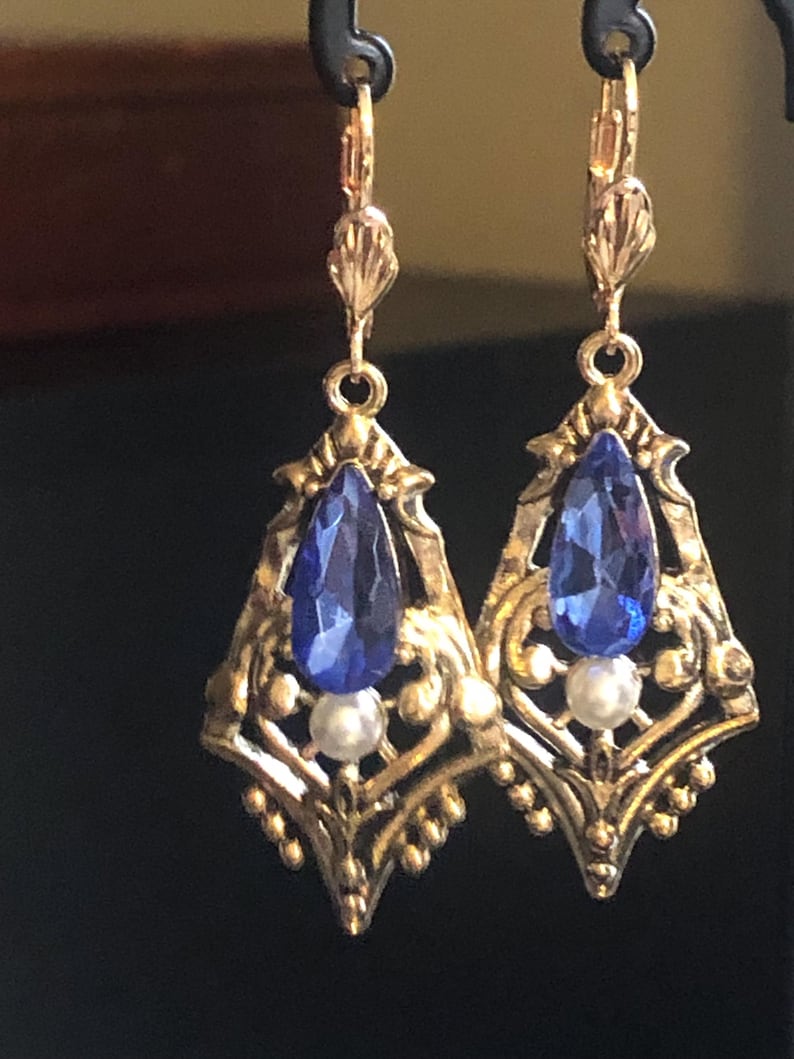 Op de afbeelding: Een paar goudkleurige filigraan oorbellen met een druppelvormige blauwe edelsteen en een kleine witte parel. De oorbellen hebben een beugel sluiting en een decoratief ontwerp.