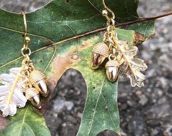 Puede incluir: Pendientes dorados de bellotas y hojas. Cada pendiente presenta tres dijes de bellota y un dije de hoja. Los pendientes se muestran sobre una hoja verde contra un fondo borroso.