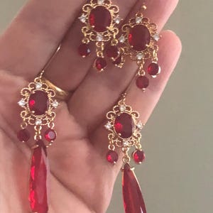 May include: Gold-toned earrings with red oval-shaped center stones and smaller red bead accents. The earrings have a floral design with clear crystal accents. One pair features a long, teardrop-shaped red stone.