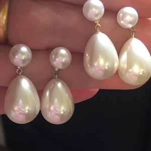 Puede incluir: Un par de pendientes de gota de perla con una perla más pequeña en la parte superior de cada pendiente. Los pendientes son de oro y tienen forma de lágrima.
