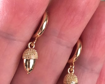 Puede incluir: Un par de pendientes de aro dorados con abalorios de bellota. Los pendientes están hechos de un metal liso y brillante y las bellotas son texturizadas.