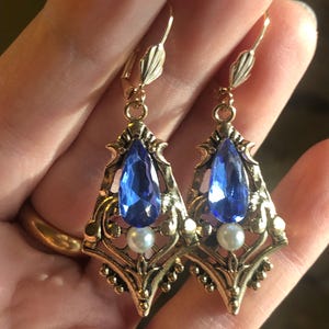 Op de afbeelding: Een paar goudkleurige oorbellen met een vintage design. Elke oorbel heeft een druppelvormige blauwe edelsteen, een kleine witte parel en sierlijke filigraan details. De oorbellen worden in een hand gehouden.