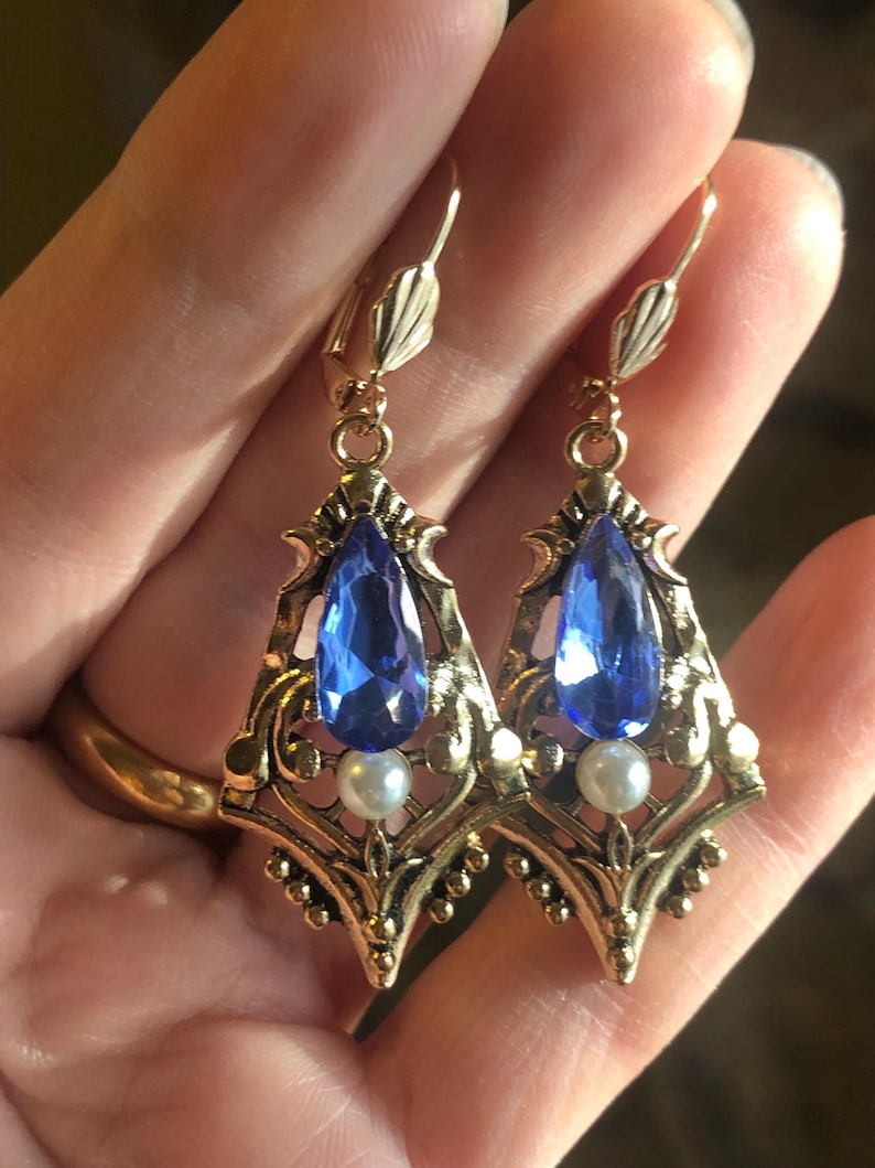 Op de afbeelding: Een paar goudkleurige oorbellen met een vintage design. Elke oorbel heeft een druppelvormige blauwe edelsteen, een kleine witte parel en sierlijke filigraan details. De oorbellen worden in een hand gehouden.