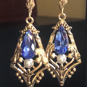 Op de afbeelding: Een paar sierlijke, goudkleurige oorbellen. Elke oorbel heeft een druppelvormige, diepblauwe edelsteen, een kleine witte parel en ingewikkelde filigraan details. De oorbellen hebben een vintage uitstraling.