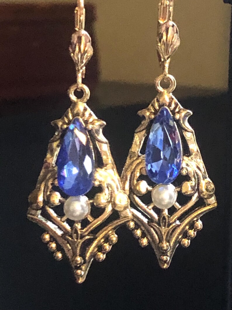 Op de afbeelding: Een paar sierlijke, goudkleurige oorbellen. Elke oorbel heeft een druppelvormige, diepblauwe edelsteen, een kleine witte parel en ingewikkelde filigraan details. De oorbellen hebben een vintage uitstraling.