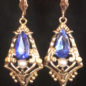 Op de afbeelding: Goudkleurige, sierlijke oorbellen met een druppelvormige blauwe edelsteen en een kleine witte parel. De oorbellen hebben een gedetailleerd filigraanontwerp en hangen aan een rode kralenketting.