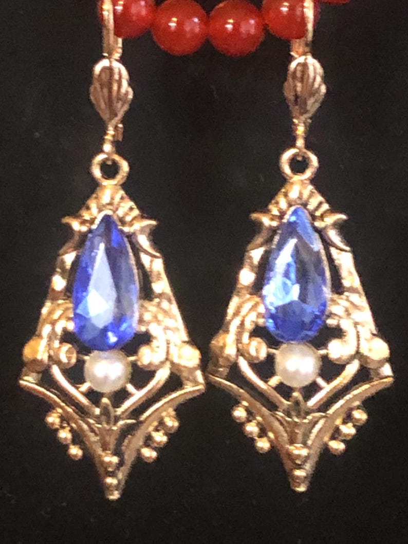 Op de afbeelding: Goudkleurige, sierlijke oorbellen met een druppelvormige blauwe edelsteen en een kleine witte parel. De oorbellen hebben een gedetailleerd filigraanontwerp en hangen aan een rode kralenketting.