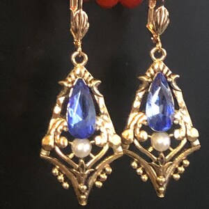 Op de afbeelding: Een paar goudkleurige, sierlijke oorbellen met een druppelvormige blauwe edelsteen en een kleine witte parel. De oorbellen hebben een filigraan ontwerp en een klapbeugel. Een rode kralenketting is zichtbaar op de achtergrond.