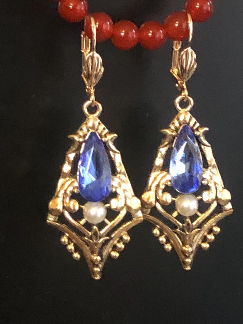 Op de afbeelding: Een paar goudkleurige, sierlijke oorbellen met een druppelvormige blauwe edelsteen en een kleine witte parel. De oorbellen hebben een filigraan ontwerp en een klapbeugel. Een rode kralenketting is zichtbaar op de achtergrond.