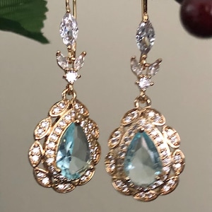 Könnte beinhalten: Ein Paar goldfarbene Ohrhänger. Jeder Ohrring hat einen tropfenförmigen, hellblauen Edelstein, der von kleinen, klaren Kristallen umgeben ist. Die Ohrringe haben ein florales Design und einen Hakenverschluss.