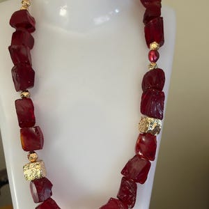 May include: A bold necklace featuring large, irregularly shaped, deep red beads. Accented with gold-colored metal elements and smaller gold beads, the necklace is displayed on a white mannequin, showcasing its striking design.