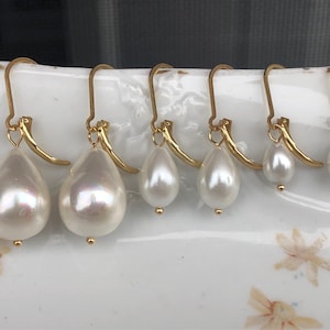 Pode incluir: Seis pares de brincos de pérolas brancas em forma de gota com ganchos dourados. As pérolas variam em tamanho, sendo a maior à esquerda e diminuindo para a direita. Os brincos são exibidos em uma superfície branca com estampa floral.