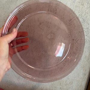May include: A clear, round, pink-tinted glass plate. The plate has a slightly raised rim with three concentric rings. The plate is held by a hand, and the background is a textured, light gray surface.