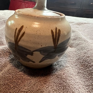 May include: A ceramic jar with a lid, featuring a beige and brown color scheme. The jar is adorned with dark blue and brown abstract leaf-like designs. The lid has a small handle. The jar is sitting on a brown towel.