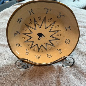 May include: A tan ceramic plate with a dark brown rim and a silver metal stand. The plate features a central sunburst design with an eye and tear, surrounded by zodiac symbols in dark blue.