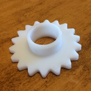May include: White plastic gear with a central hole. The gear has 16 teeth.