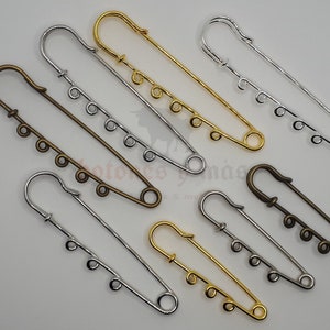 Może przedstawiać: Sześć szpilek bezpieczeństwa w różnych kolorach, w tym złoty, srebrny i brązowy. Każda szpilka ma ozdobną pętlę na końcu i szereg mniejszych pętli wzdłuż długości szpilki.