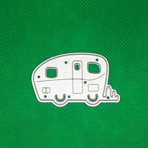 May include: A white metal die cut of a camper trailer with rounded edges and two windows. The camper has a door and a small window on the side. The camper is on a green background.