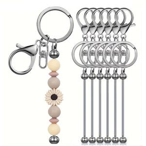 Op de afbeelding: Een verzameling sleutelhangers met zilverkleurige hardware. Eén sleutelhanger heeft een decoratief kralenontwerp met crème, roze en bruine kralen en een kleine bloem. De andere sleutelhangers hebben een eenvoudig ontwerp met een ring en een lange, dunne metalen staaf.