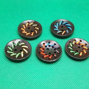 Op de afbeelding: Vijf houten knopen met kleurrijke draadborduursels. Elke knop heeft een andere kleurcombinatie van draad, waaronder groen, oranje, blauw en bruin.