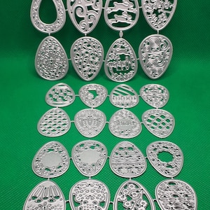 May include: A set of 20 metal die-cut Easter egg shapes. The eggs are in various designs, including floral, geometric, and bunny patterns. The eggs are silver in colour and are arranged in rows on a green background.