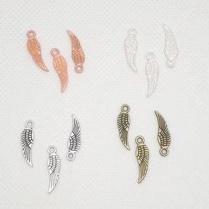 Könnte beinhalten: Ein Set mit sechs Metall-Engelsflügel-Anhängern in drei verschiedenen Ausführungen: Gold, Silber und Antik-Bronze. Jeder Anhänger verfügt über ein detailliertes Flügeldesign mit einer Öse zum Anbringen an Schmuck oder anderen Bastelarbeiten.