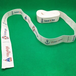 Könnte beinhalten: Weißes Baumwollband mit nautischen Motiven. Das Band zeigt einen Leuchtturm, einen Anker, einen Rettungsring und den Text "Land & Sea", "Ocean Lover" und "Vintage Voyage".