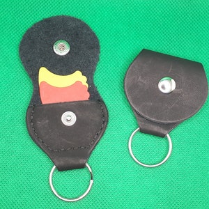 Könnte beinhalten: Zwei schwarze Leder-Schlüsselanhänger mit Metallringen. Ein Schlüsselanhänger hat eine Klappe, die sich öffnet und einen kleinen roten und gelben Gitarren-Plektron enthüllt.