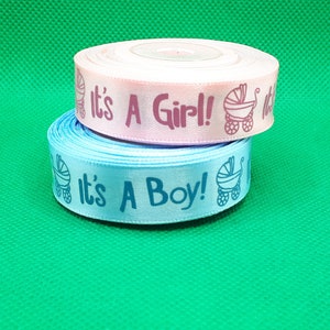 Könnte beinhalten: Zwei Rollen Satinband, eine rosa und eine blaue, mit dem Text "It's A Girl!" und "It's A Boy!" bedruckt. Jedes Band zeigt ein kleines Bild eines Kinderwagens.