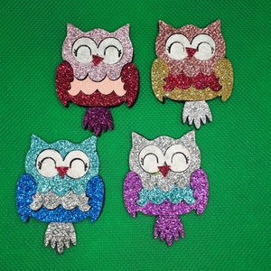 Könnte beinhalten: Vier glitzernde Eulenschmuckstücke in verschiedenen Farben. Die Eulen sind rosa, gold, blau und lila. Sie haben weiße Augen und einen roten Schnabel.