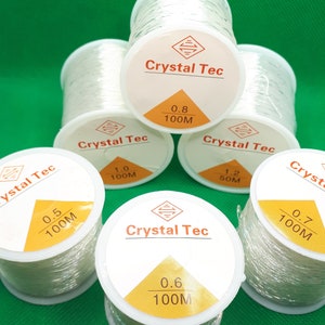 May include: Six spools of clear elastic thread, each with a yellow label that reads "Crystal Tec" and indicates the thickness and length of the thread. The spools are arranged in a circle on a green surface.