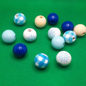 Op de afbeelding: Een verzameling houten kralen in verschillende kleuren en patronen. De kralen omvatten effen blauwe, lichtblauwe en natuurlijke houttinten. Sommige kralen hebben een blauw-wit ruitpatroon of een sneeuwvlokontwerp. De kralen liggen op een groene ondergrond.