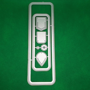 Puede incluir: Un troquel de metal plateado con varias formas, incluyendo un rectángulo, un cuadrado, un círculo y una lágrima. El troquel está sobre un fondo verde.