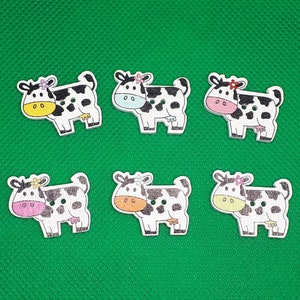 Könnte beinhalten: Sechs hölzerne Kuhknöpfe mit schwarzen und weißen Flecken. Jede Kuh hat eine Blume in einer anderen Farbe auf dem Kopf. Die Kühe schauen alle nach rechts.