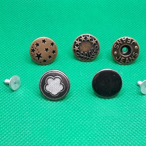 May include: Assortment of six metal jean buttons in various designs, including stars, leaves, and a plain black button. Two silver metal button fasteners are also included.
