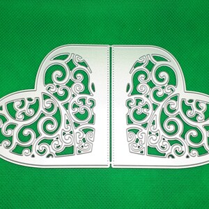 Op de afbeelding: Een hartvormig, gestanst papieren ambacht met ingewikkelde, wervelende filigraanpatronen. De twee helften zijn in het midden verbonden, waardoor een symmetrisch ontwerp ontstaat. Het ambacht is gemaakt van een lichtgekleurd materiaal tegen een groene achtergrond.