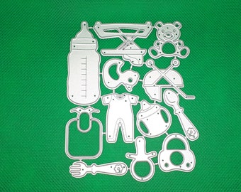 May include: A collection of silver-colored baby-themed die cuts, including a baby bottle, bib, onesie, pacifier, spoon, and teddy bear. The die cuts are arranged on a green background.