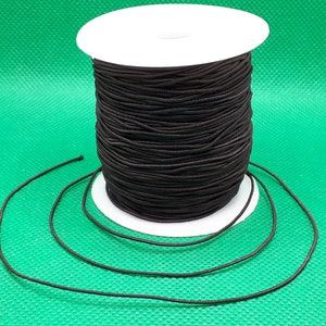 Può includere: Una bobina di cordino elastico nero. Il cordino è avvolto attorno a una bobina di cartone bianca. Una lunghezza del cordino si estende dalla bobina, appoggiata su una superficie verde. Questo cordino elastico è adatto a vari progetti di artigianato e cucito.