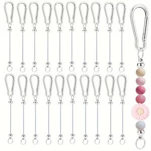 Op de afbeelding: Een verzameling zilverkleurige karabijnhaakclips met bevestigde draad en kleine metalen kralen. Eén clip heeft een decoratieve bedel met houten en roze kralen, en een roze bloem. De clips zijn ontworpen voor diverse toepassingen.