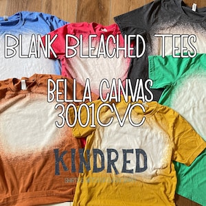 Puede incluir: Una colección de seis camisetas blancas blanqueadas en varios colores, incluyendo azul, rojo, gris, verde, naranja y amarillo. Las camisetas están dispuestas sobre una superficie de madera y presentan un diseño blanqueado blanco. El texto "BLANK BLEACHED TEES" está escrito en negro en la parte superior de la imagen. El texto "BELLA CANVAS 3001 CVC" está escrito en negro debajo de las camisetas. El texto "KINDRED" está escrito en negro en la parte inferior de la imagen.
