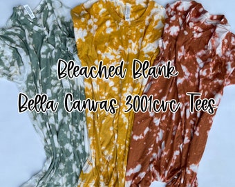 May include: Three bleached blank Bella Canvas 3001 CVC t-shirts in different colours: green, yellow, and orange. The shirts have a tie-dye pattern.