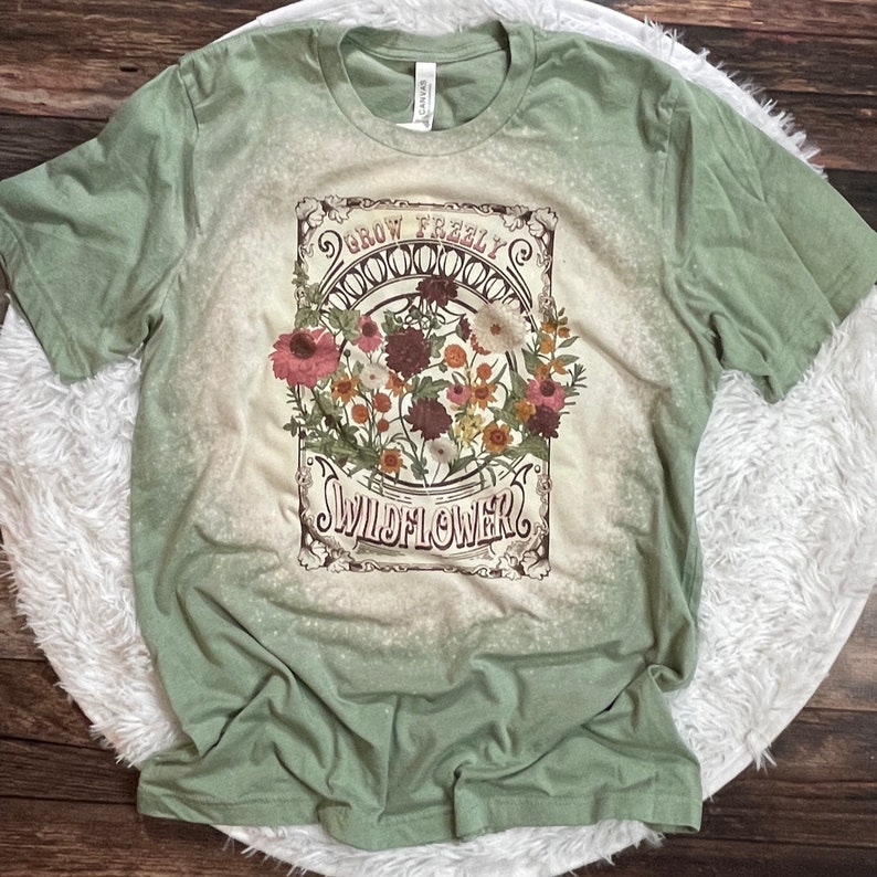 K&ouml;nnte beinhalten: Gr&uuml;nes T-Shirt mit einem Vintage-Grafikdruck von Blumen und dem Text "Grow Freely Wildflower".