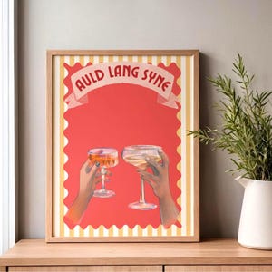 Puede incluir: Una impresión artística enmarcada con dos manos brindando con copas de champán. La obra de arte tiene un fondo rojo con las palabras "Auld Lang Syne" en una pancarta. El marco es de color madera clara y la impresión tiene un borde a rayas amarillas y blancas.