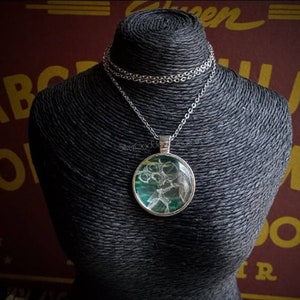 May include: A silver-toned pendant necklace with a round, glass-covered charm. The charm features a green and white abstract design. The necklace is displayed on a black mannequin against a dark red background.