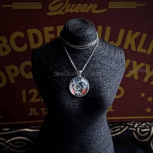 May include: A silver-toned pendant necklace with a round, glass-covered charm. The charm features a colorful, detailed design. The necklace is displayed on a black mannequin against a backdrop of a Ouija board. The text "Silver Goddess Creations" is visible.